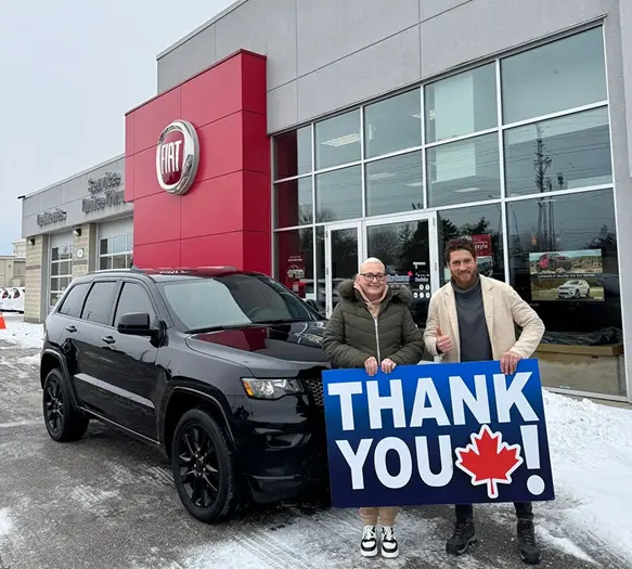 A happy couple holds a large THANK YOU! sign in front of a black SUV at a Fiat dealership on a snowy day.