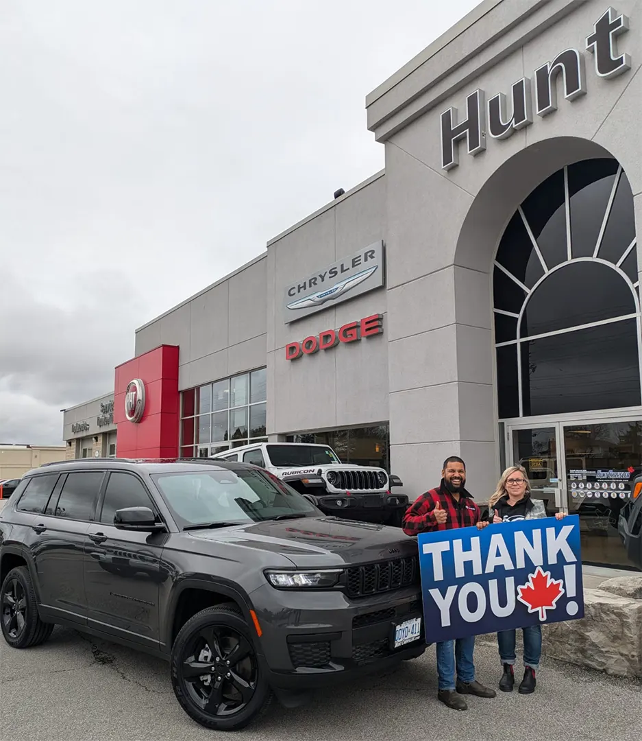 A black SUV parked outside a dealership, with two individuals holding a large sign that says THANK YOU! in front of a Chrysler Dodge store.