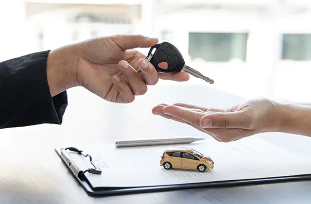 A person hands over car keys to another, beside a clipboard and a small toy car, symbolizing a vehicle purchase or transfer.