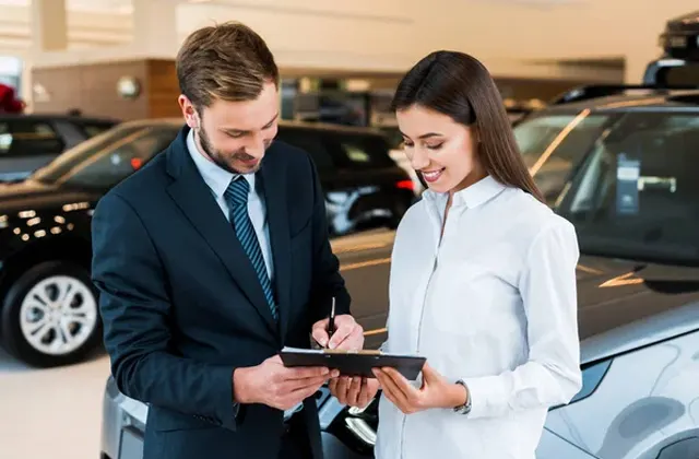 A man in a suit discusses details with a woman in a white blouse, both examining a tablet in a car dealership filled with vehicles.
