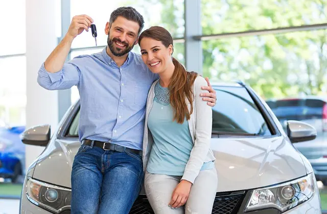 A happy couple poses together on a car in a dealership, with one holding car keys and smiling. Bright, spacious showroom in the background.