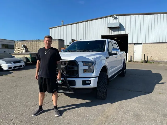 A person stands confidently next to a lifted white truck in a parking lot, with an industrial building in the background under a clear blue sky.