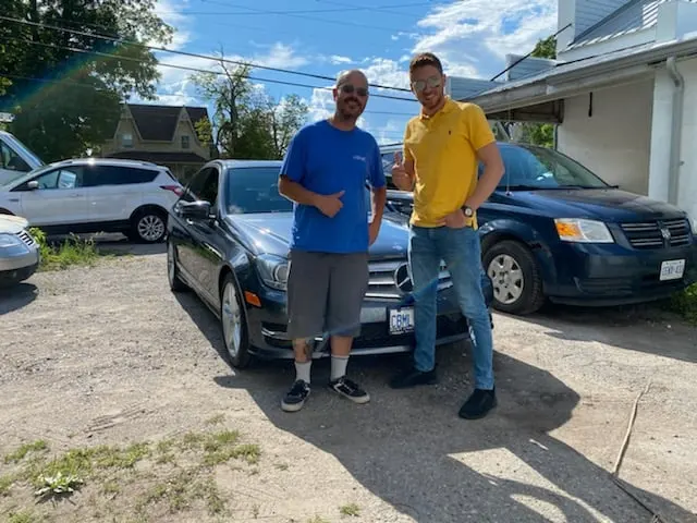 Two men stand beside a black Mercedes-Benz in a gravel parking area, both giving thumbs up. Various cars are parked in the background.
