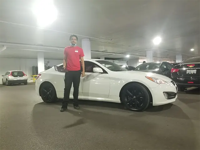 A person stands next to a white sports car with black wheels in a dimly lit parking garage. Other vehicles are parked nearby.
