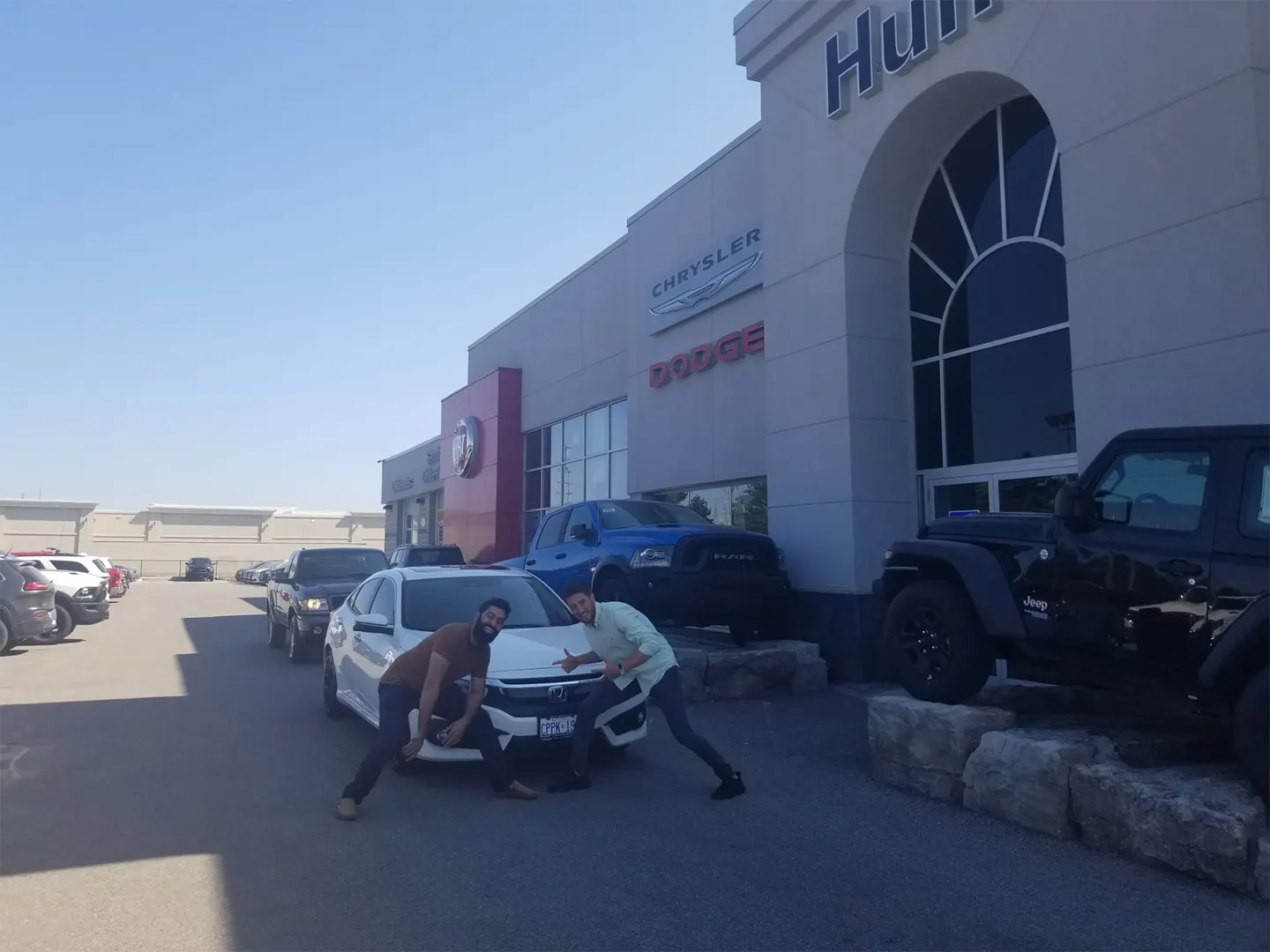 Two people playfully pose in front of a white SUV at a car dealership, with various vehicles parked in the background.